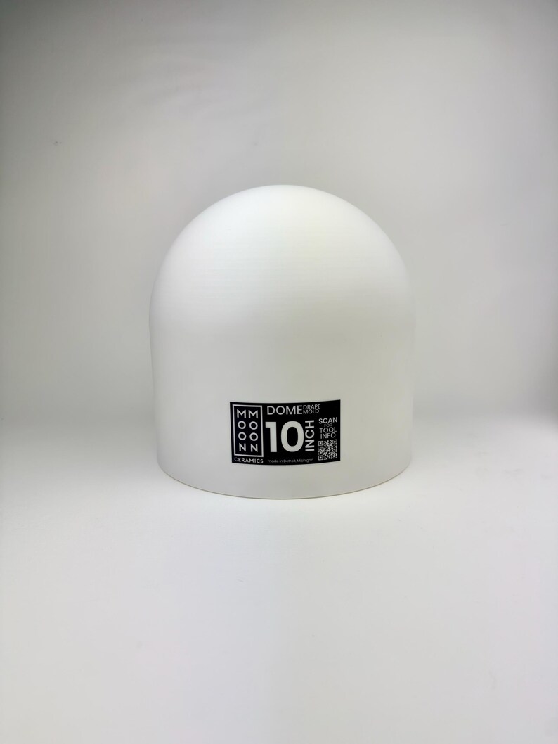 May include: A white dome drape mold with a smooth, matte surface. A black rectangular label on the front displays the text Moon Ceramics, Dome Drape Mold, 10 Inch, and includes a QR code for tool information. The object has a rounded top.