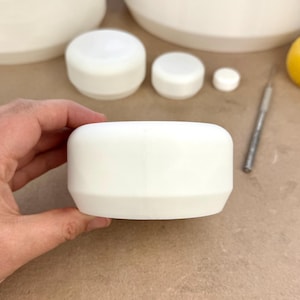 Könnte beinhalten: Eine Sammlung weißer Keramikbehälter und Deckel. Der Fokus liegt auf einem weißen, zylindrischen Behälter mit abgerundeter Oberseite und einem kleineren Deckel. Ein Bastelmesser ist im Hintergrund zu sehen.
