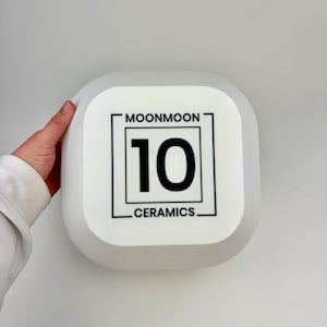 May include: A square, white, translucent ceramic object with rounded corners. The center features a black graphic design consisting of a square border, the text MOONMOON at the top, the number 10 in the middle, and CERAMICS at the bottom.