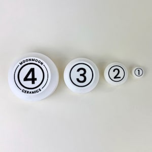 May include: Four white ceramic round lids with black numbers 1, 2, 3, and 4. The lid with the number 4 also has the text "MOONMOON CERAMICS" around the edge.