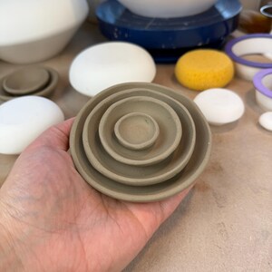 May include: A set of four nested circular clay dishes in a matte taupe finish. The dishes are stacked concentrically, with the smallest dish in the center and the largest on the outside, featuring smooth, rounded edges and a simple, shallow bowl shape.