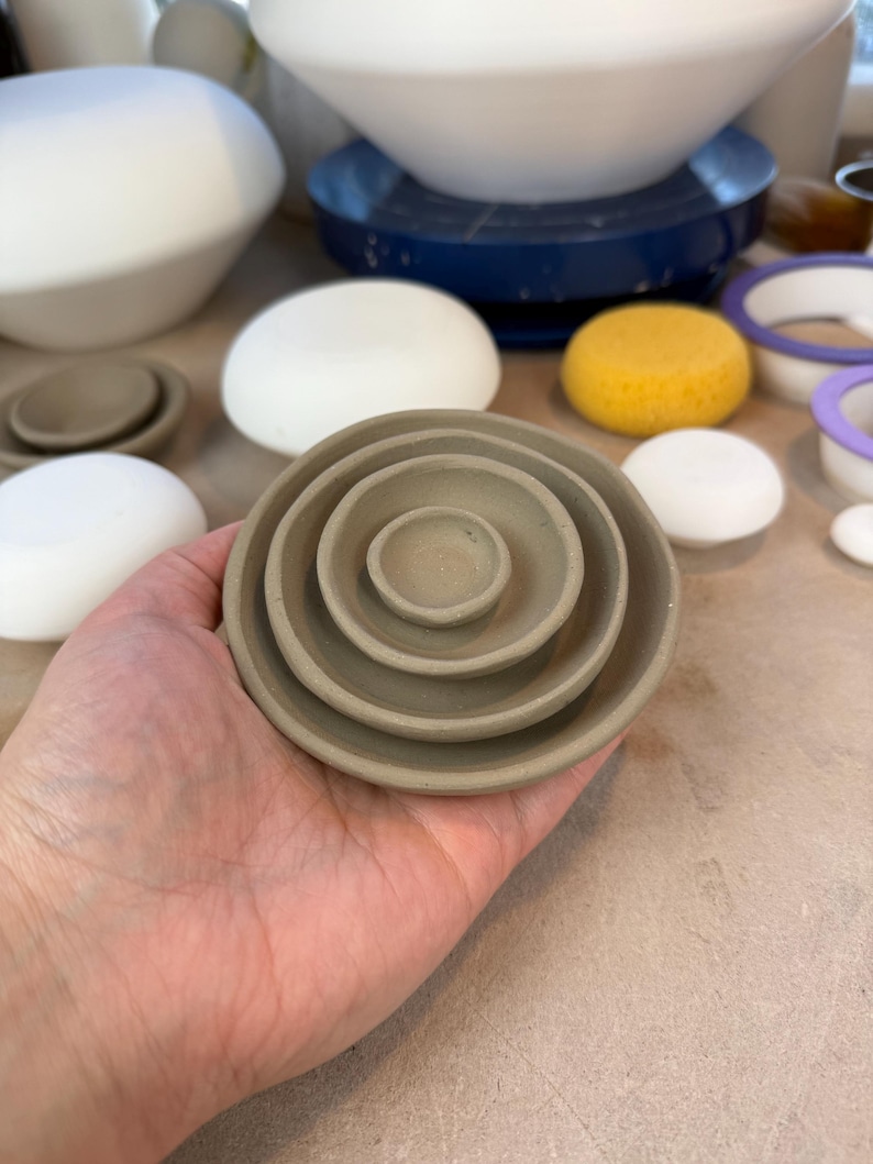 May include: A set of four nested circular clay dishes in a matte taupe finish. The dishes are stacked concentrically, with the smallest dish in the center and the largest on the outside, featuring smooth, rounded edges and a simple, shallow bowl shape.