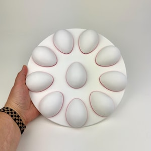 Puede incluir: Un soporte blanco para huevos con un diseño circular, que contiene nueve huevos blancos. El soporte tiene un contorno rojo alrededor de cada huevo. El soporte está siendo sostenido por una mano.