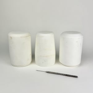 Puede incluir: Tres recipientes cilíndricos blancos con tapas redondeadas están dispuestos en fila. Una fina herramienta de metal con punta se encuentra delante de ellos. Los objetos están sobre un fondo blanco.