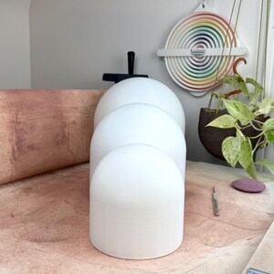 May include: Three white, smooth, dome-shaped objects of varying sizes arranged in a vertical line. Each object has a cylindrical base and a rounded top, featuring a matte finish. The objects are positioned on a textured, light brown surface.