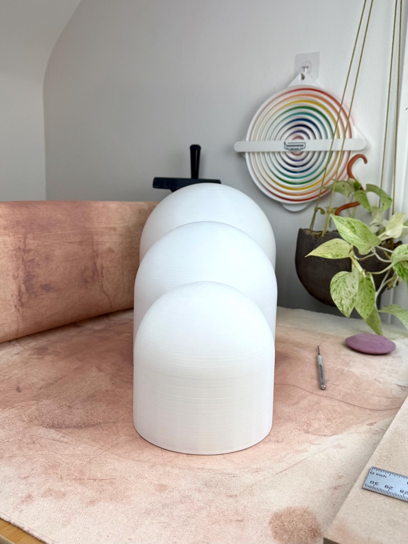 May include: Three white, smooth, dome-shaped objects of varying sizes arranged in a vertical line. Each object has a cylindrical base and a rounded top, featuring a matte finish. The objects are positioned on a textured, light brown surface.