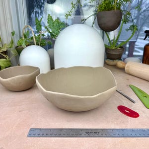 May include: Two beige ceramic bowls with irregular, wavy rims. A metal ruler is positioned in the foreground, showing a length of 30 centimeters. The bowls are placed on a light-colored surface alongside a wooden rolling pin and plastic tools.