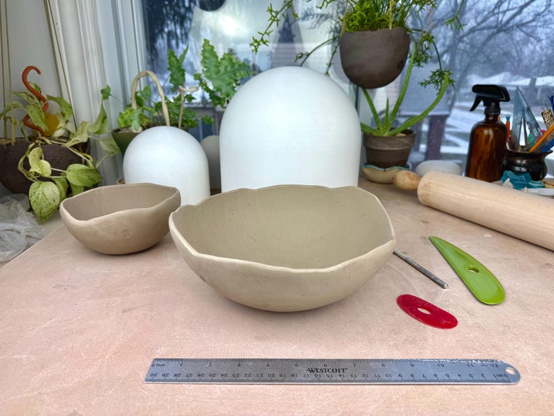 May include: Two beige ceramic bowls with irregular, wavy rims. A metal ruler is positioned in the foreground, showing a length of 30 centimeters. The bowls are placed on a light-colored surface alongside a wooden rolling pin and plastic tools.