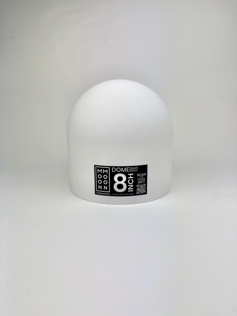 May include: A white 8 inch dome drape mold by Moon Ceramics. The object is a smooth, semi-spherical tool with a flat base. A black label on the front displays the text Moon Ceramics, Dome Drape Mold, 8 Inch, and includes a QR code for tool info.