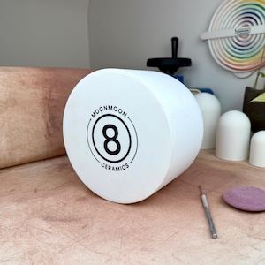 May include: A cylindrical white ceramic object featuring a black circular logo on the front face. The logo displays the number 8 in the center, surrounded by the text MOONMOON CERAMICS. The object has a smooth, matte finish and a flat circular front.