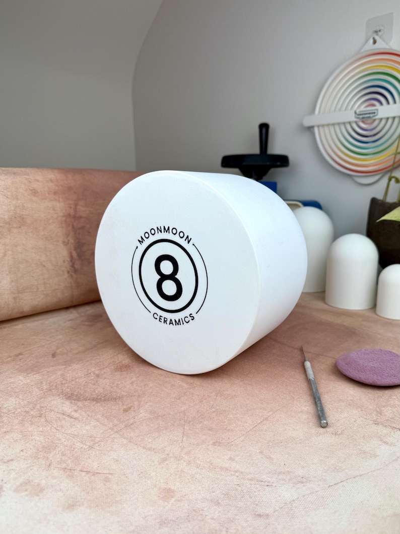 May include: A cylindrical white ceramic object featuring a black circular logo on the front face. The logo displays the number 8 in the center, surrounded by the text MOONMOON CERAMICS. The object has a smooth, matte finish and a flat circular front.