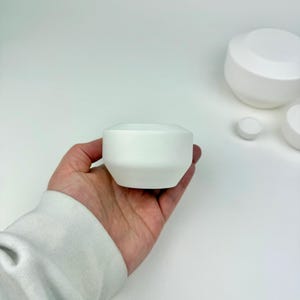 May include: A small, matte white ceramic bowl with a faceted, geometric shape. The bowl features a wide, angled rim that tapers down to a narrower base, presenting a clean, solid white surface without any patterns or additional decorative details.