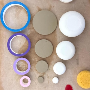 May include: A collection of crafting supplies including four circular rolls of tape in blue, purple, lavender, and light pink, four solid tan circular discs, four white circular objects of varying sizes, a yellow sponge, and a red plastic scraper.
