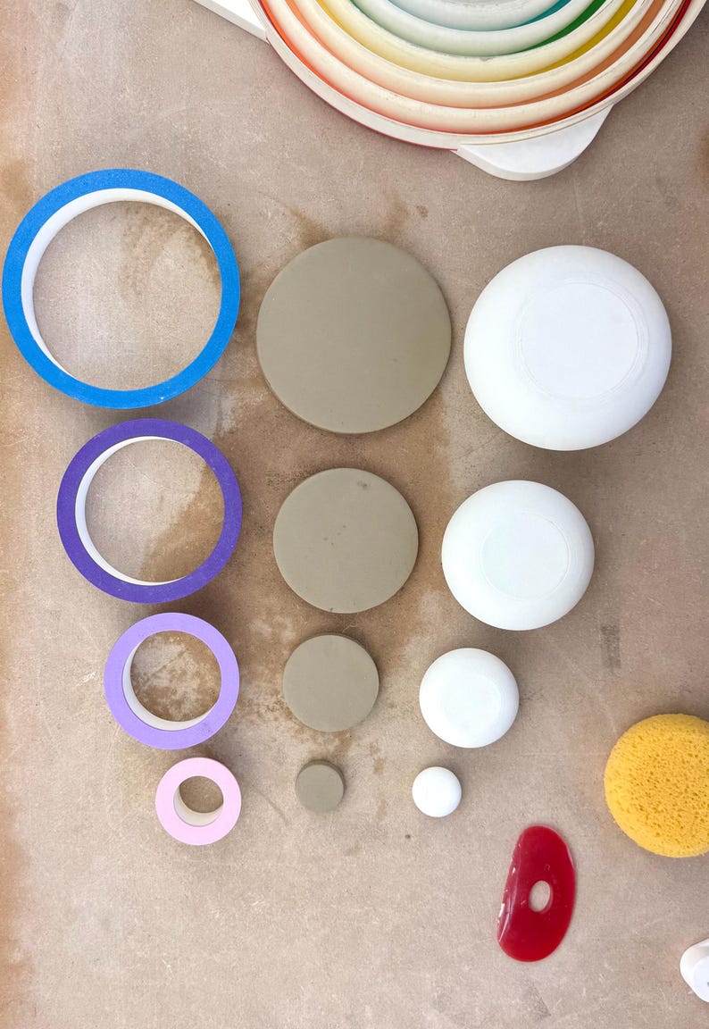 May include: A collection of crafting supplies including four circular rolls of tape in blue, purple, lavender, and light pink, four solid tan circular discs, four white circular objects of varying sizes, a yellow sponge, and a red plastic scraper.