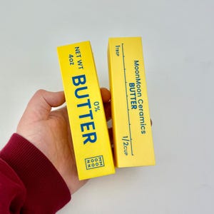 Può includere: Due bastoncini di burro in ceramica gialla con testo blu. Uno recita "BUTTER" a grandi lettere, "NET WT 4oz" e "0%" sul davanti. L'altro recita "MoonMoon Ceramics BUTTER" e presenta segni di misurazione per 1 cucchiaio e 1/2 tazza.