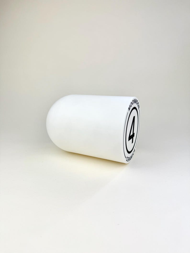 Op de afbeelding: Een wit keramisch object met afgeronde uiteinden. Het object heeft een zwart cirkelvormig logo met het nummer 4 erin en de woorden "MOONMOON CERAMICS" rond de cirkel. Het object staat op een witte achtergrond.