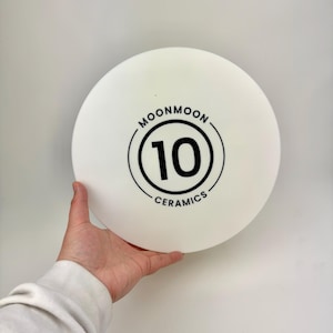 May include: A circular, matte white ceramic plate featuring a black graphic design in the center. The design consists of the number 10 enclosed within a double circle, with the text MOONMOON CERAMICS printed along the top and bottom curves.