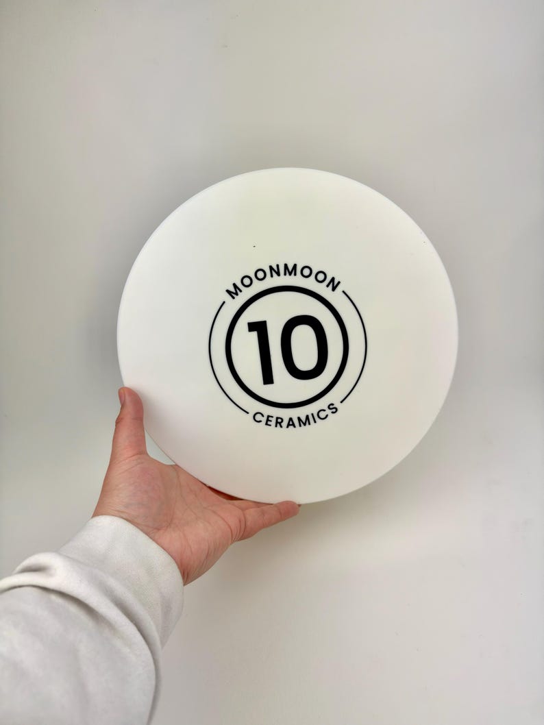 May include: A circular, matte white ceramic plate featuring a black graphic design in the center. The design consists of the number 10 enclosed within a double circle, with the text MOONMOON CERAMICS printed along the top and bottom curves.