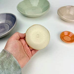 May include: A small, round, speckled cream-colored ceramic bowl with a rectangular maker's mark stamped on the bottom. Surrounding it are four other ceramic bowls in varying sizes and colors, including light green, purple, beige, and orange.