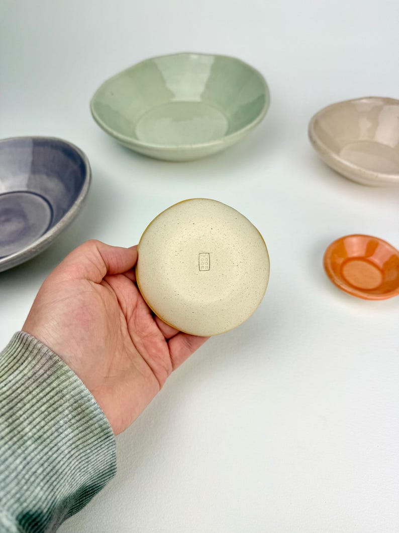 May include: A small, round, speckled cream-colored ceramic bowl with a rectangular maker's mark stamped on the bottom. Surrounding it are four other ceramic bowls in varying sizes and colors, including light green, purple, beige, and orange.