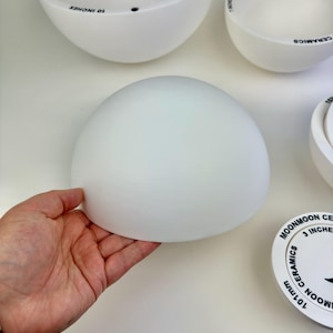 May include: A set of white ceramic hemispherical bowls of varying sizes. One bowl is held, showing a smooth, matte finish. Other bowls are visible with printed text including 'MOONMOON CERAMICS' and measurements in inches and millimeters.
