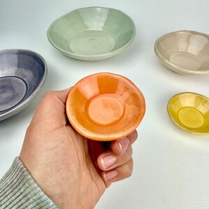 May include: A collection of five small, round, ceramic bowls with a glossy finish. The bowls are colored in light green, beige, purple, bright yellow, and a vibrant orange, which is held in the foreground, showcasing a smooth, glazed surface.