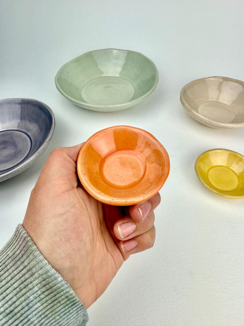 May include: A collection of five small, round, ceramic bowls with a glossy finish. The bowls are colored in light green, beige, purple, bright yellow, and a vibrant orange, which is held in the foreground, showcasing a smooth, glazed surface.