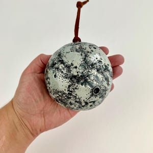Op de afbeelding: Een bespikkelde keramische bol met een leren lus. De bol is voornamelijk wit met zwarte en grijze spikkels. Hij heeft twee kleine gaatjes en een bruine leren lus om op te hangen.
