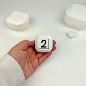 May include: A white square-shaped button with rounded corners featuring a black number 2 printed inside a thin black square border. The button is held in a palm, with two additional white square objects visible in the background.
