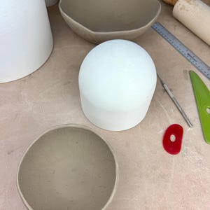 Op de afbeelding: Een verzameling keramische potten in verschillende stadia van voltooiing. De stukken omvatten een grote witte koepel, een lichtbruine kom en een grotere, onregelmatig gevormde kom. Pottenbakkersgereedschap, waaronder een liniaal en een rood vormgereedschap, zijn ook zichtbaar.