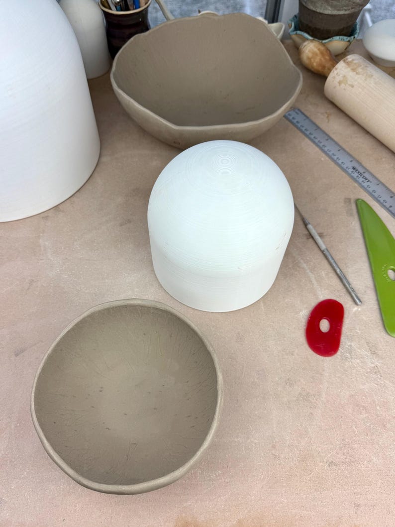 Op de afbeelding: Een verzameling keramische potten in verschillende stadia van voltooiing. De stukken omvatten een grote witte koepel, een lichtbruine kom en een grotere, onregelmatig gevormde kom. Pottenbakkersgereedschap, waaronder een liniaal en een rood vormgereedschap, zijn ook zichtbaar.