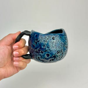 Op de afbeelding: Een handgemaakte keramische mok met een uniek, getextureerd oppervlak. De mok heeft een diepblauwe glazuur met donkerblauwe en zwarte accenten en een zwart handvat. Het ontwerp lijkt op een hemellichaam. De mok wordt in een hand gehouden.