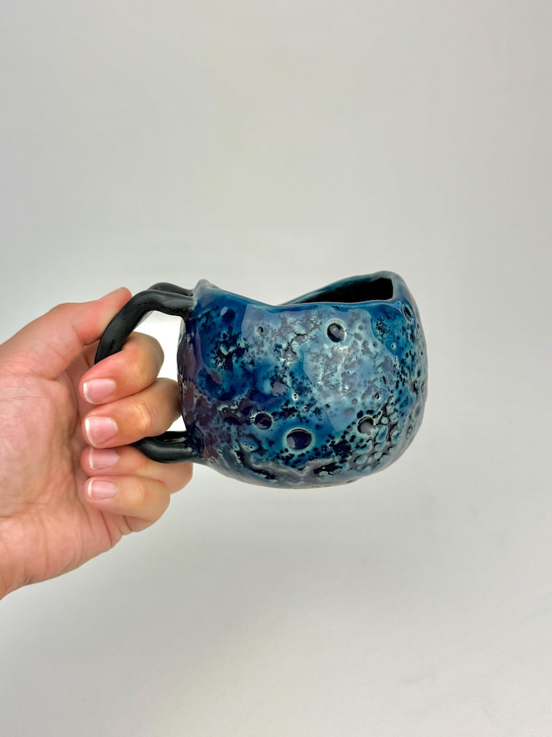 Op de afbeelding: Een handgemaakte keramische mok met een uniek, getextureerd oppervlak. De mok heeft een diepblauwe glazuur met donkerblauwe en zwarte accenten en een zwart handvat. Het ontwerp lijkt op een hemellichaam. De mok wordt in een hand gehouden.