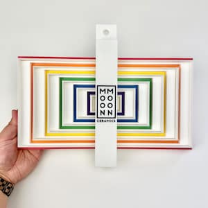 May include: A set of nested rectangular ceramic trays in a rainbow color gradient, featuring red, orange, yellow, green, blue, and purple borders. A vertical white strap across the center displays the black text MOON MOON CERAMICS.