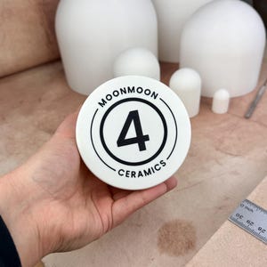 Op de afbeelding: Een wit keramisch stuk met de tekst "MOONMOON 4 CERAMICS" in een zwart cirkelvormig ontwerp. Op de achtergrond staan ​​verschillende witte keramische vormen van verschillende afmetingen. Een liniaal is rechts zichtbaar.