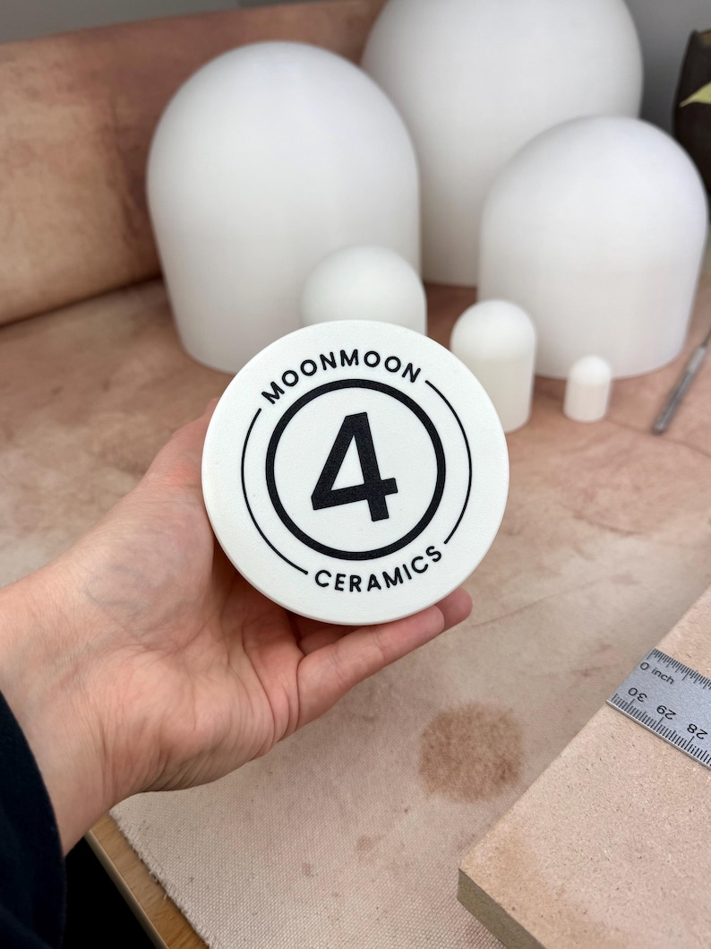 Op de afbeelding: Een wit keramisch stuk met de tekst "MOONMOON 4 CERAMICS" in een zwart cirkelvormig ontwerp. Op de achtergrond staan ​​verschillende witte keramische vormen van verschillende afmetingen. Een liniaal is rechts zichtbaar.