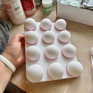 May include: A white tray holds twelve white, spherical objects with a red base. The tray is held by a person wearing a white wristband. Jars of pink and white paint are in the background.