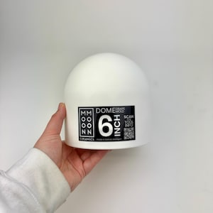 May include: A white ceramic dome drape mold with a black label featuring the text MOON CERAMICS, DOME DRAPE MOLD, 6 INCH, SCAN FOR TOOL INFO, and a QR code. The mold has a smooth, rounded top surface and a flat base, designed for pottery shaping.