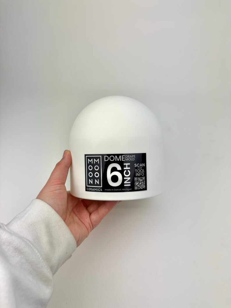 May include: A white ceramic dome drape mold with a black label featuring the text MOON CERAMICS, DOME DRAPE MOLD, 6 INCH, SCAN FOR TOOL INFO, and a QR code. The mold has a smooth, rounded top surface and a flat base, designed for pottery shaping.