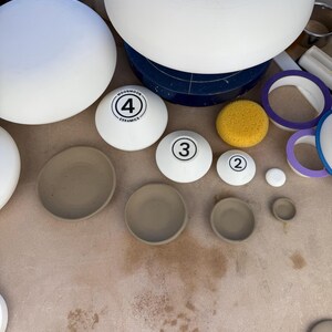 May include: A collection of white ceramic pottery forms and beige clay bowls. Several white rounded pieces are marked with numbers 2, 3, and 4, with the text MOONMOON CERAMICS visible on the largest numbered piece. A yellow sponge is included.