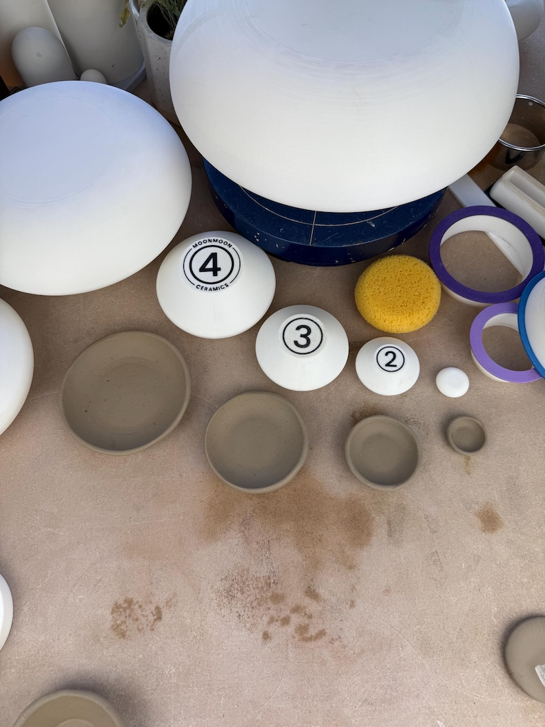 May include: A collection of white ceramic pottery forms and beige clay bowls. Several white rounded pieces are marked with numbers 2, 3, and 4, with the text MOONMOON CERAMICS visible on the largest numbered piece. A yellow sponge is included.