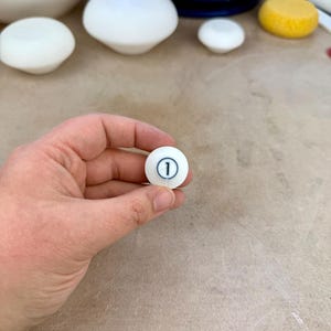 May include: A small, spherical white ceramic object featuring a black circular outline with the number 1 printed in the center. The surface of the object is smooth and matte, and it is held in a position that displays the front-facing numerical detail.