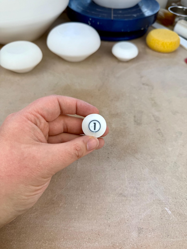 May include: A small, spherical white ceramic object featuring a black circular outline with the number 1 printed in the center. The surface of the object is smooth and matte, and it is held in a position that displays the front-facing numerical detail.