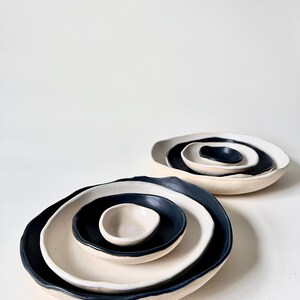 May include: Two sets of three nested ceramic bowls with irregular, organic edges. Each set features a contrasting color scheme with matte black interiors and off-white exteriors, arranged in a stack from largest to smallest bowl.