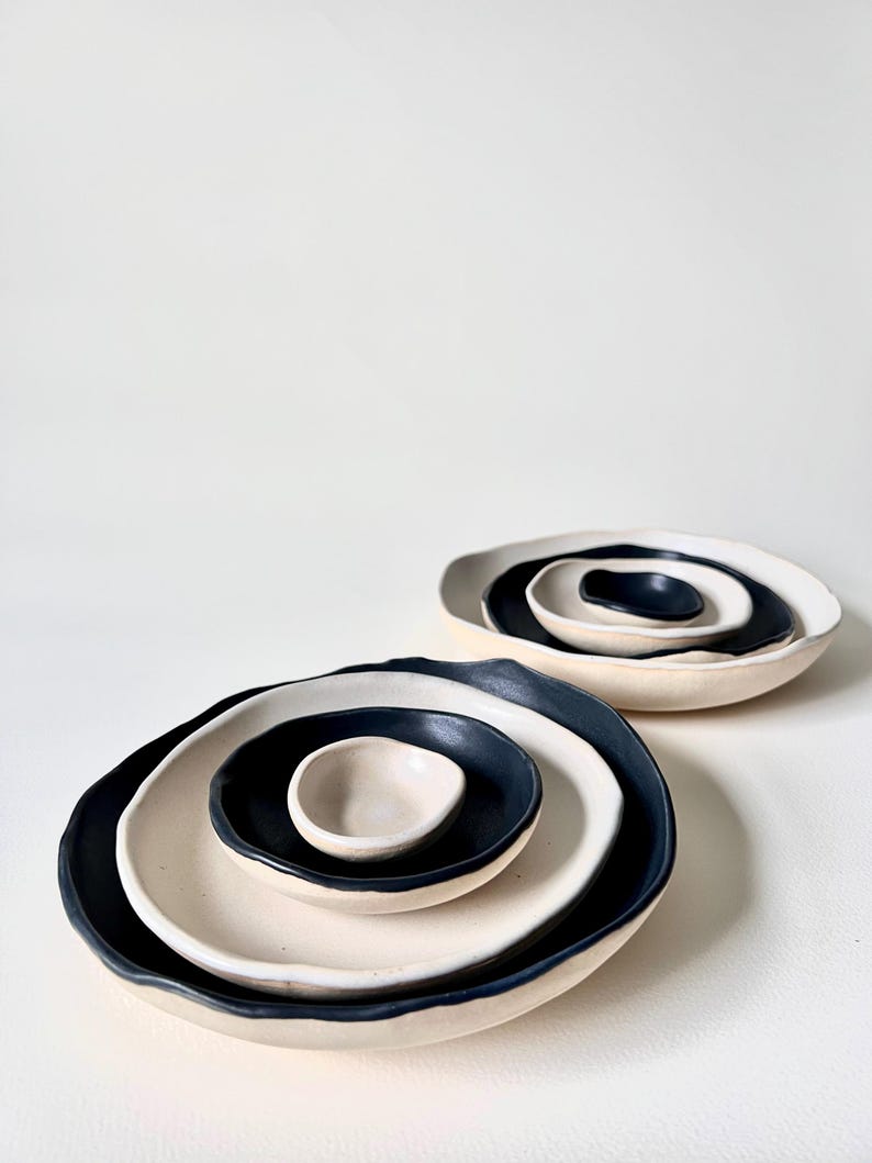 May include: Two sets of three nested ceramic bowls with irregular, organic edges. Each set features a contrasting color scheme with matte black interiors and off-white exteriors, arranged in a stack from largest to smallest bowl.