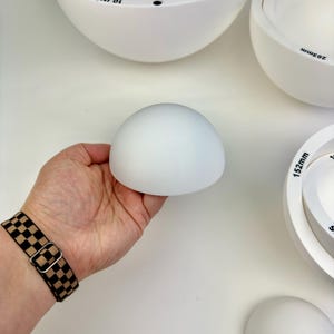 May include: A set of white ceramic hemispherical molds for pottery. The molds are various sizes, with some featuring printed text such as 10 inches, 7 inches, 203mm, and 152mm. The smooth, matte white surfaces are designed for shaping clay forms.