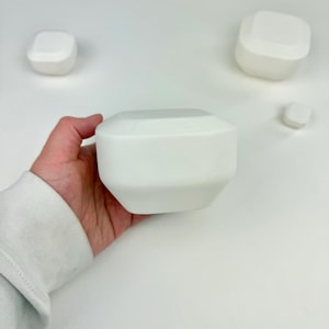 May include: A set of four white, matte-finished geometric objects with faceted edges. The objects vary in size, with one large piece held in the palm, two medium-sized pieces, and one small piece, all featuring a smooth, solid, and opaque surface.
