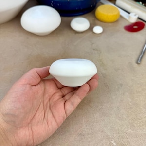 May include: A white ceramic piece with a rounded top and flared base is held in a hand. Other white ceramic items of different sizes are in the background, along with a yellow disc and a blue pottery wheel.
