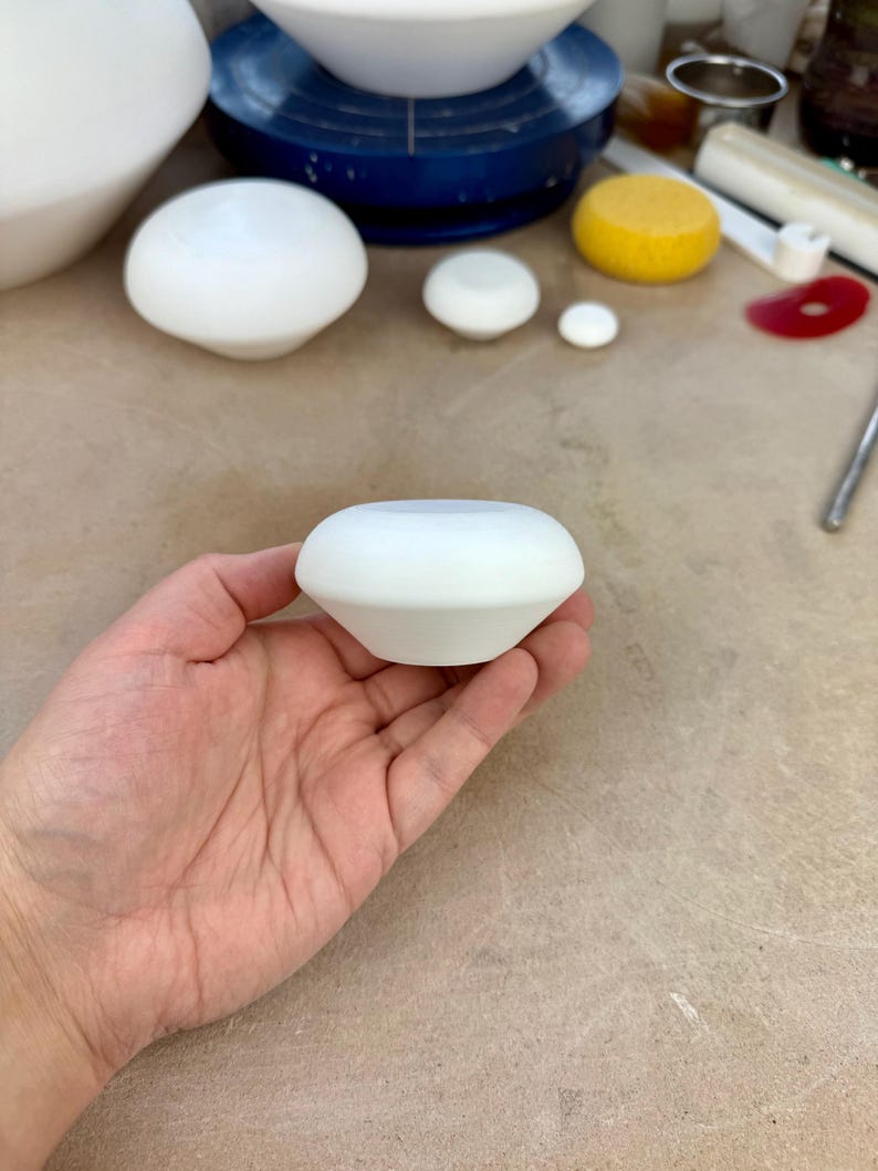 May include: A small, matte white ceramic object with a rounded, tapered shape. The piece features a smooth, uniform surface and a wide, flat top opening, showcasing a clean, geometric form that fits comfortably within the palm of a hand.