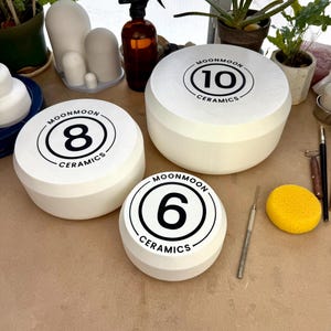 May include: Three white ceramic bowls of varying sizes, each featuring a black circular logo on the top surface. The logos display the text MOONMOON CERAMICS surrounding the numbers 6, 8, and 10 respectively. The bowls have a smooth, matte finish.